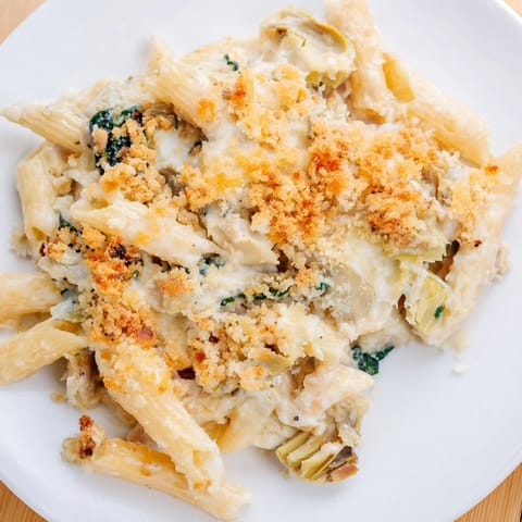A spoon serves a generous portion of creamy Artichoke Pasta Bake onto a white plate, revealing tender spinach and artichokes.