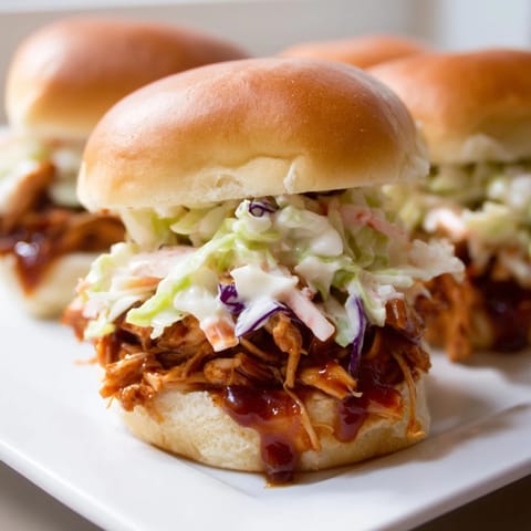A close-up of tender pulled BBQ Chicken Sliders, garnished with tangy slaw and fresh herbs.  