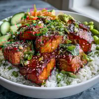 Golden-brown broiled salmon cubes sit atop fluffy white rice in this Baked Salmon Rice Bowl, garnished with crisp cucumber and shredded carrots.